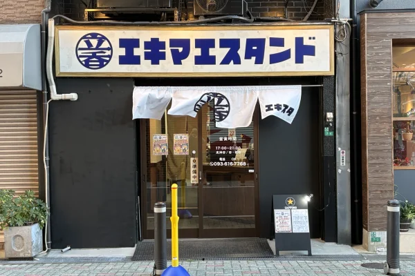 白地に紺の文字で「エキマエスタンド」と書かれた看板と、「エキスタ」ののれんがかかる入口