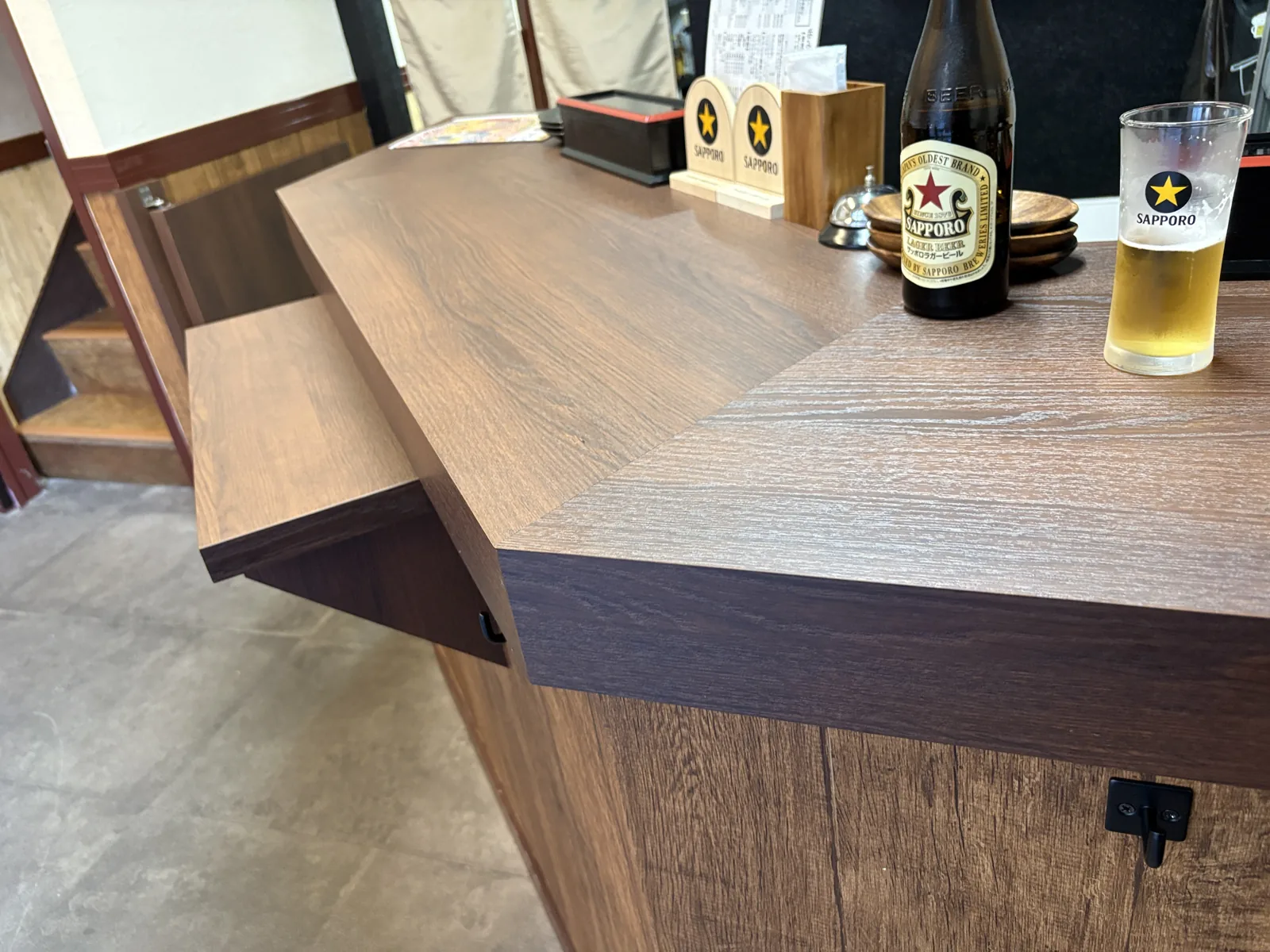 立ち飲みカウンターの角。赤星の中瓶とサッポロのグラス。カウンターの側面に荷物置きの小さな棚