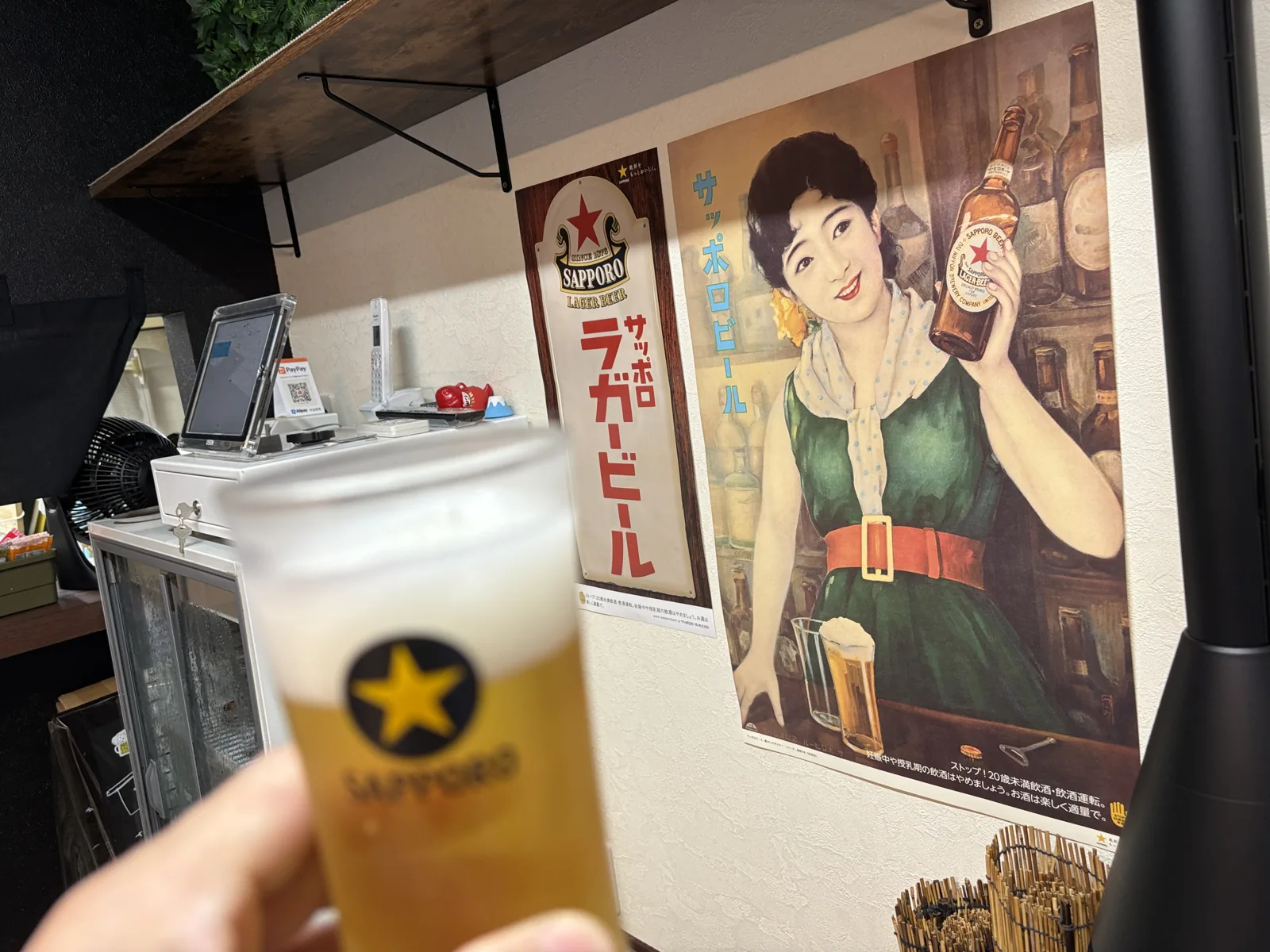 赤星を注いだグラスを掲げる。背後にサッポロラガービールの看板と、瓶を持つ女性の美人画ポスター