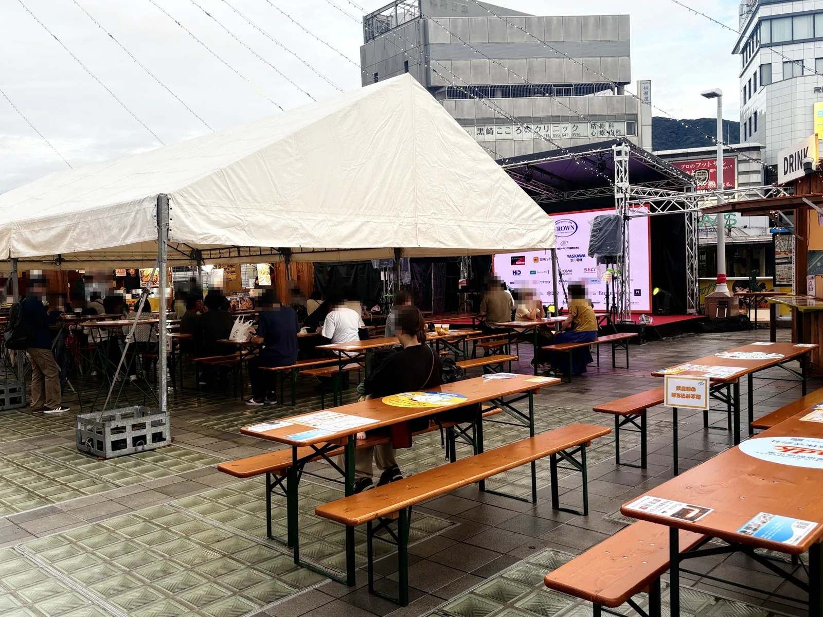 雨あがりのステージ前。白いテントの横に木製テーブルとベンチが並び、空席が目立つ
