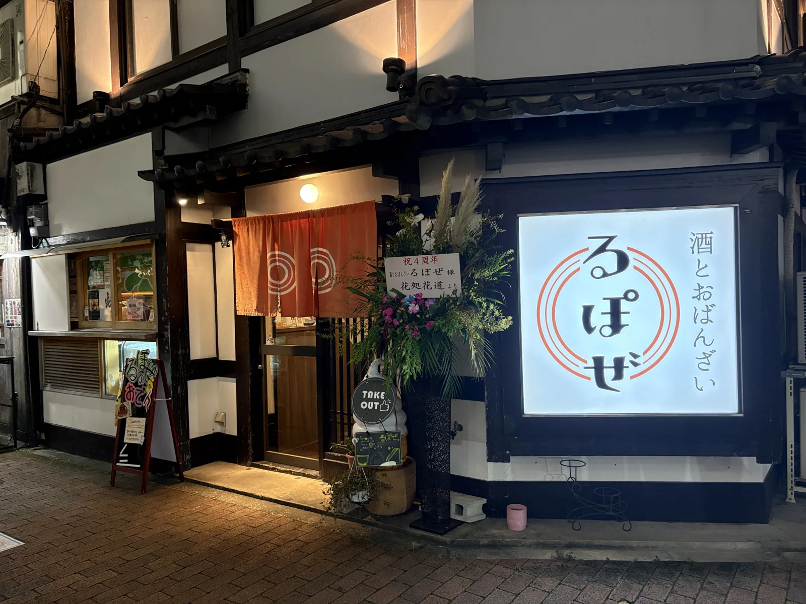 白壁に黒い柱、瓦の庇の古民家。オレンジののれんと「酒とおばんざい るぽぜ」の光る看板