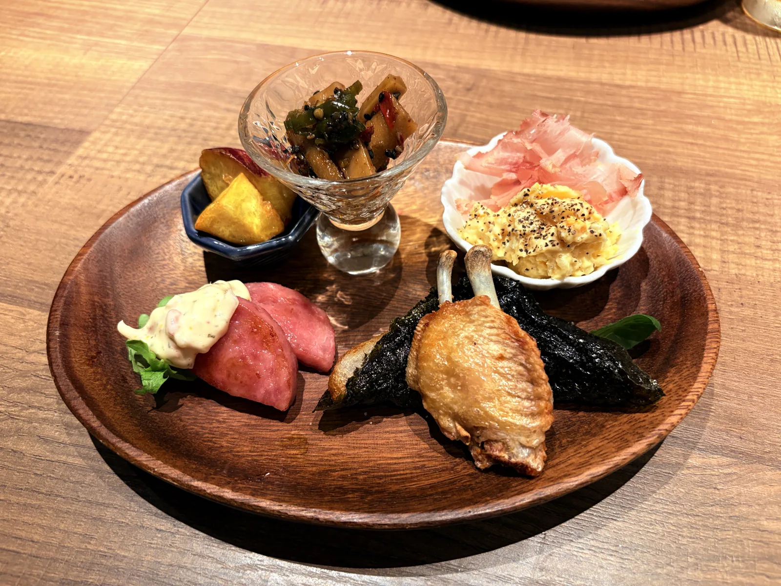 木のプレートに手羽の一夜干し、とろろ磯辺揚げ、雲仙ハムのソテー、ぴりからごぼう、ポテトサラダ、さつまいも