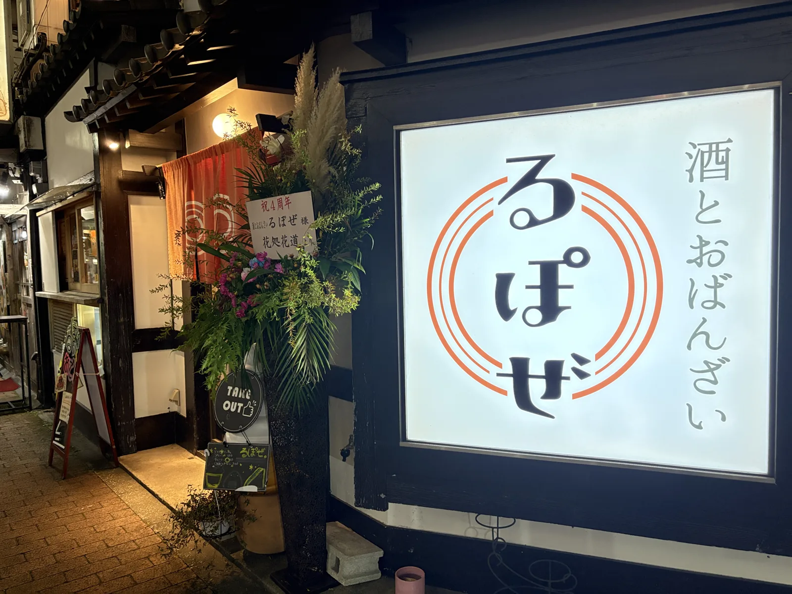 夜の「酒とおばんざい るぽぜ」の外観。祝4周年のスタンド花とTAKE OUTの看板