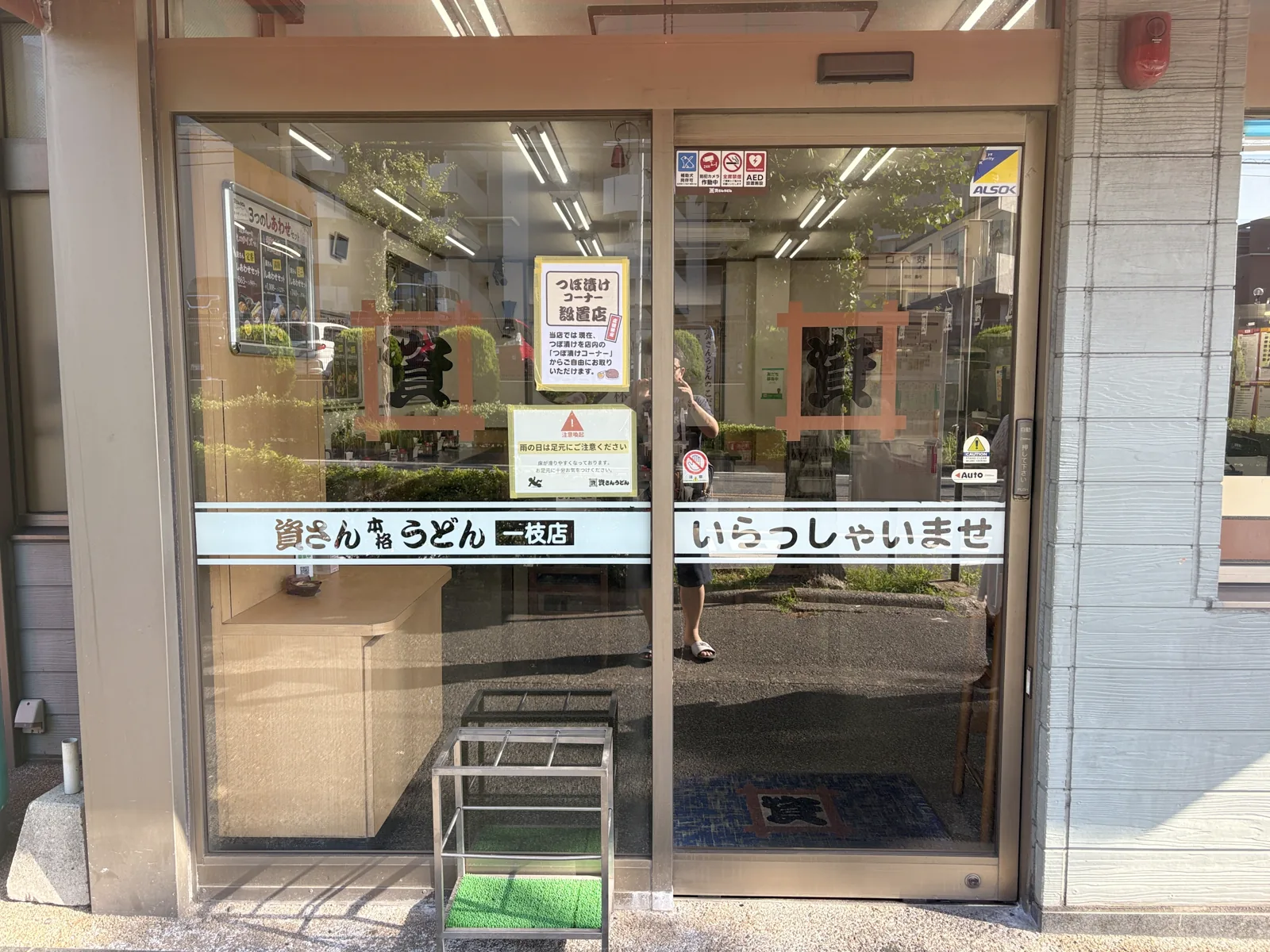 入口のガラス戸。「資さん本格うどん 一枝店」「いらっしゃいませ」の文字と「つぼ漬けコーナー設置店」の貼り紙