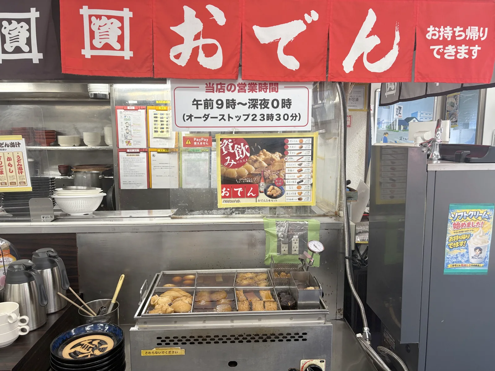 赤い「おでん」ののれんの下におでんの鍋。「当店の営業時間 午前9時〜深夜0時」の掲示と「資飲みのススメ」のポスター