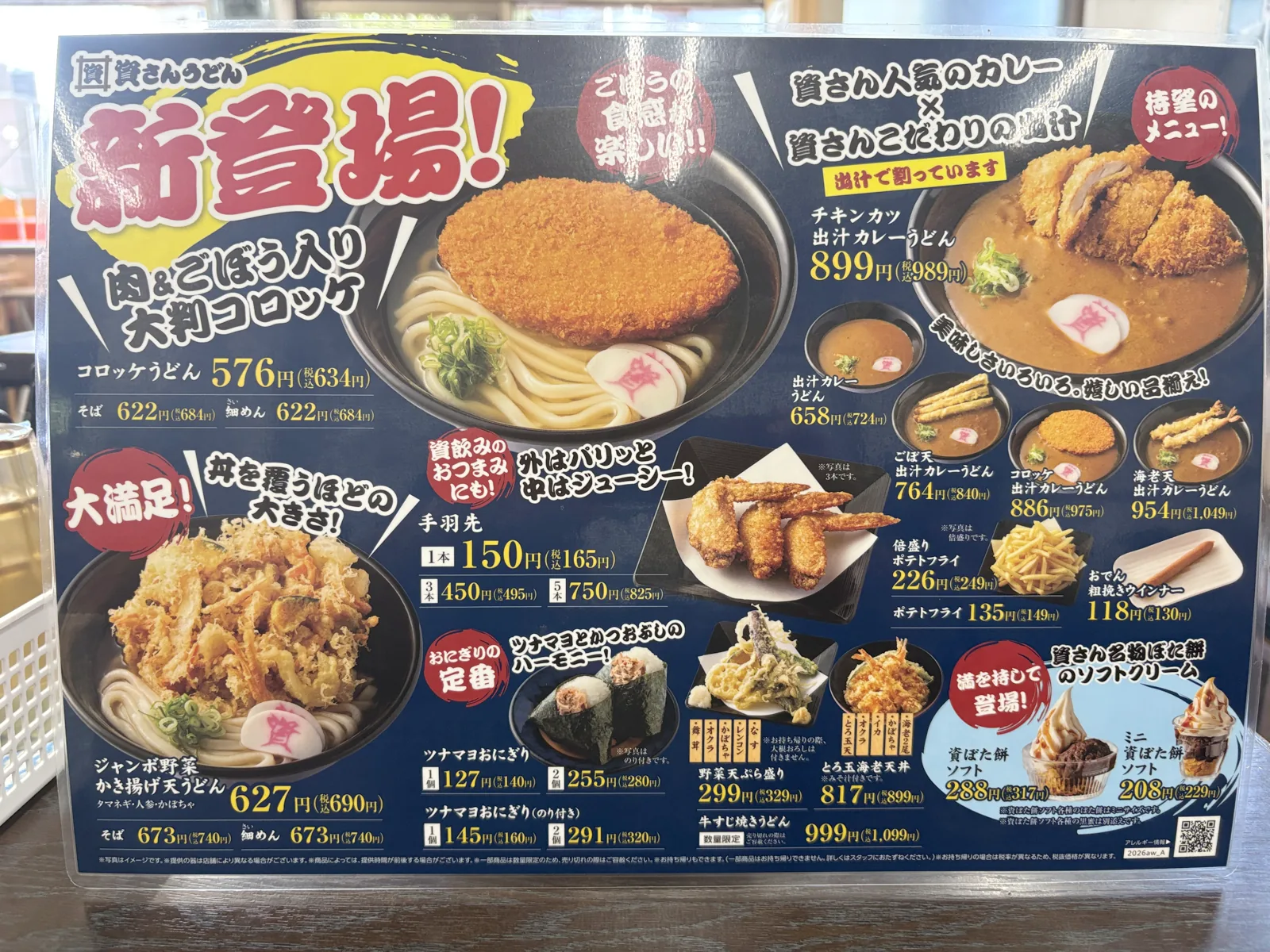 資さんうどんの店頭POP「新登場!」。コロッケうどん、出汁カレーうどん、ジャンボ野菜かき揚げ天うどん、手羽先、資ぼた餅ソフトの価格が並ぶ