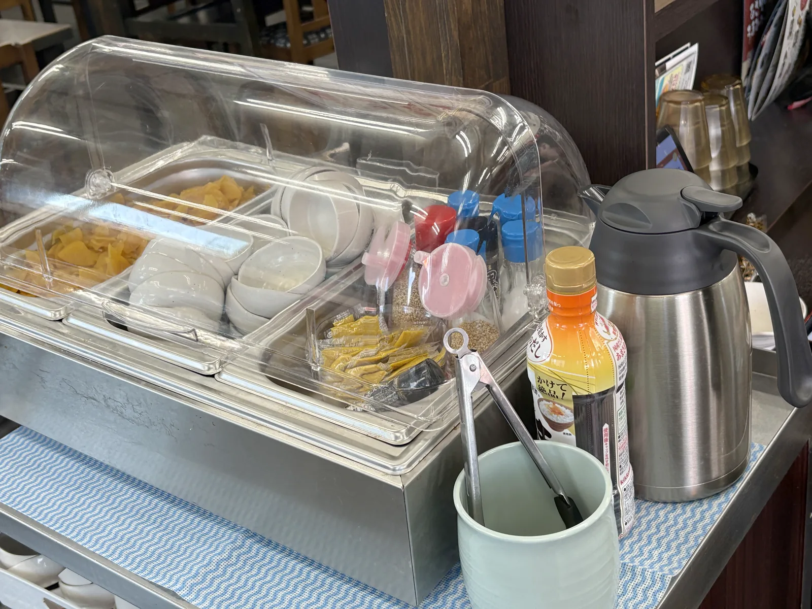 陣山店の薬味コーナー。クリアカバーの中につぼ漬けと小皿、調味料が並び、横にポット