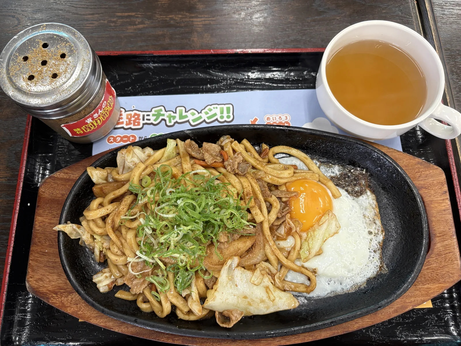 鉄板の焼きうどんに半熟の目玉焼きとネギ。右上にはカップに入った出汁