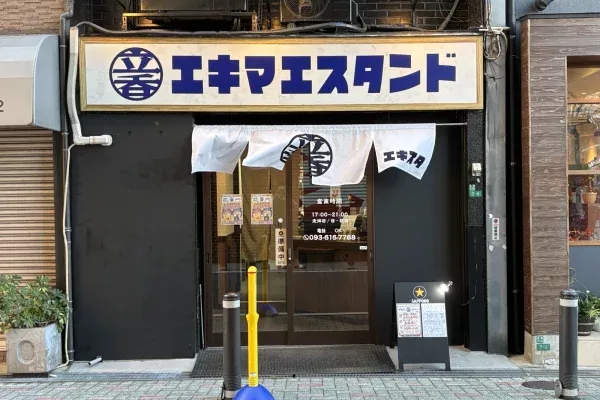 白地に紺の文字で「エキマエスタンド」と書かれた看板と、「エキスタ」ののれんがかかる入口