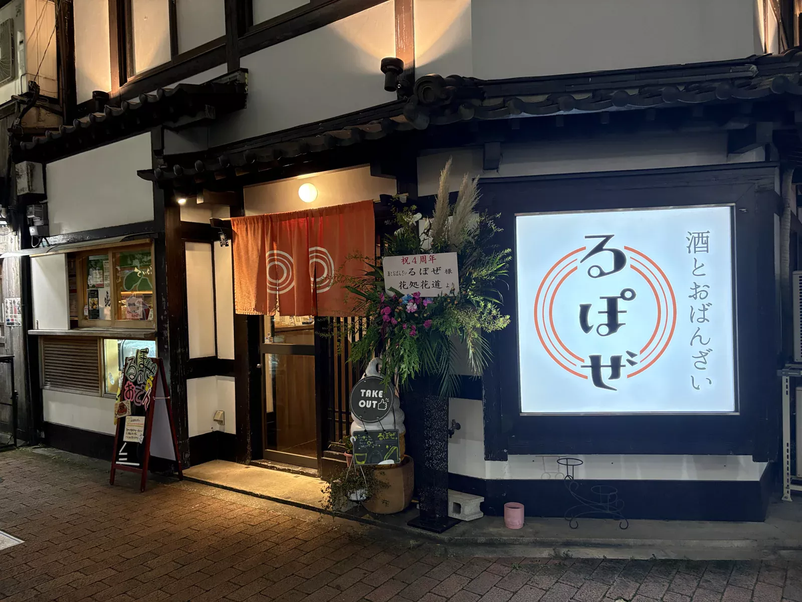 白壁に黒い柱、瓦の庇の古民家。オレンジののれんと「酒とおばんざい るぽぜ」の光る看板