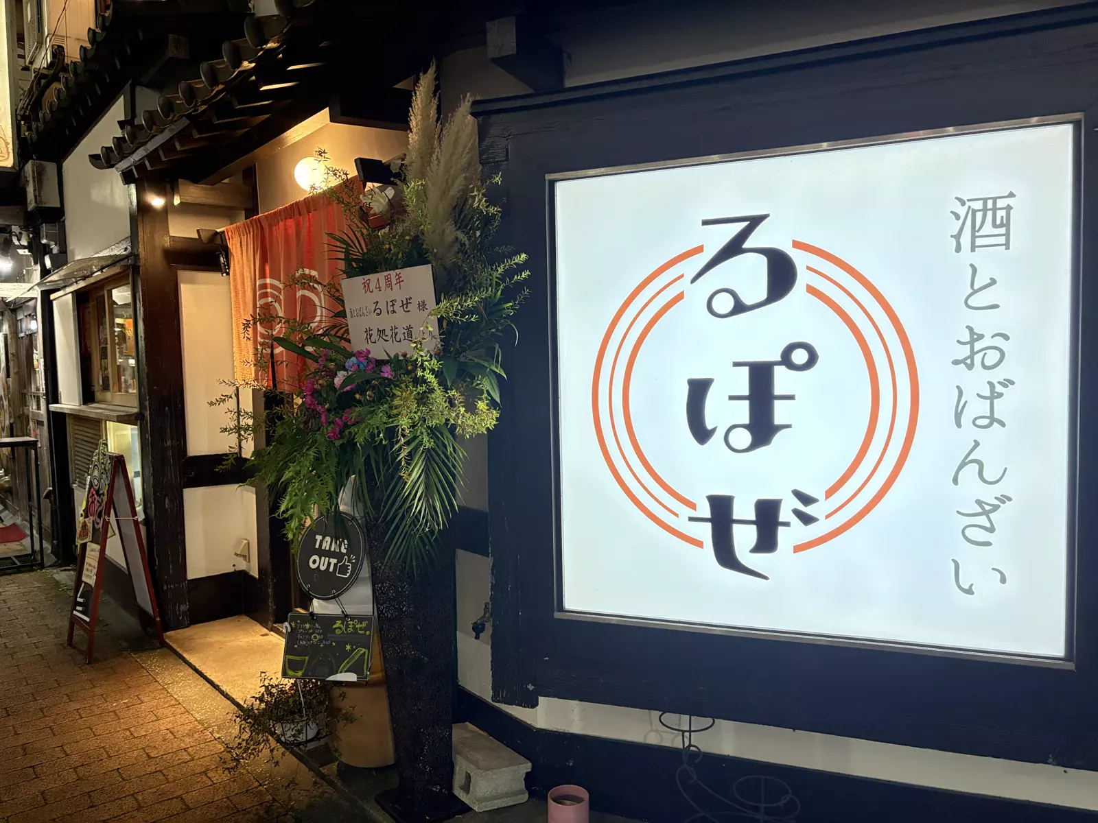 夜の「酒とおばんざい るぽぜ」の外観。祝4周年のスタンド花とTAKE OUTの看板