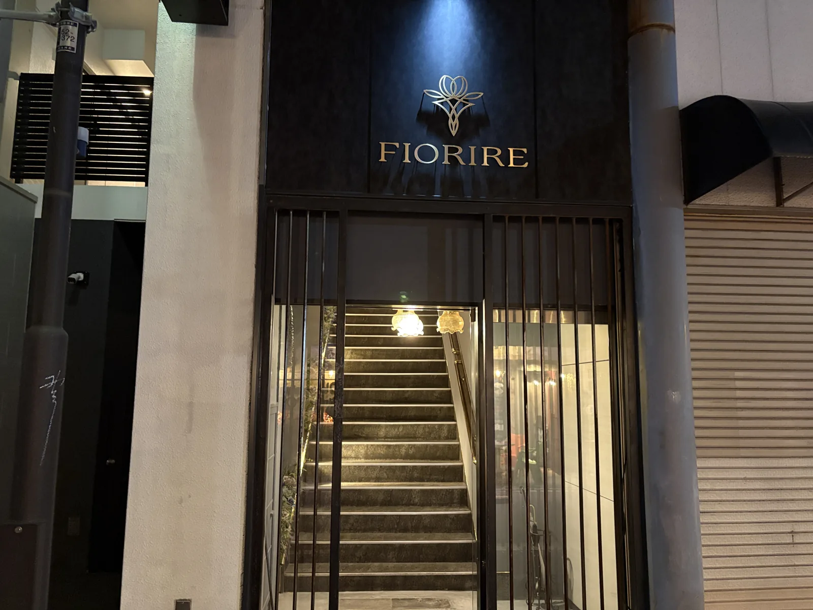 黒い壁に金色の花のロゴと「FIORIRE」の文字。格子の奥にランプに照らされた階段が続く