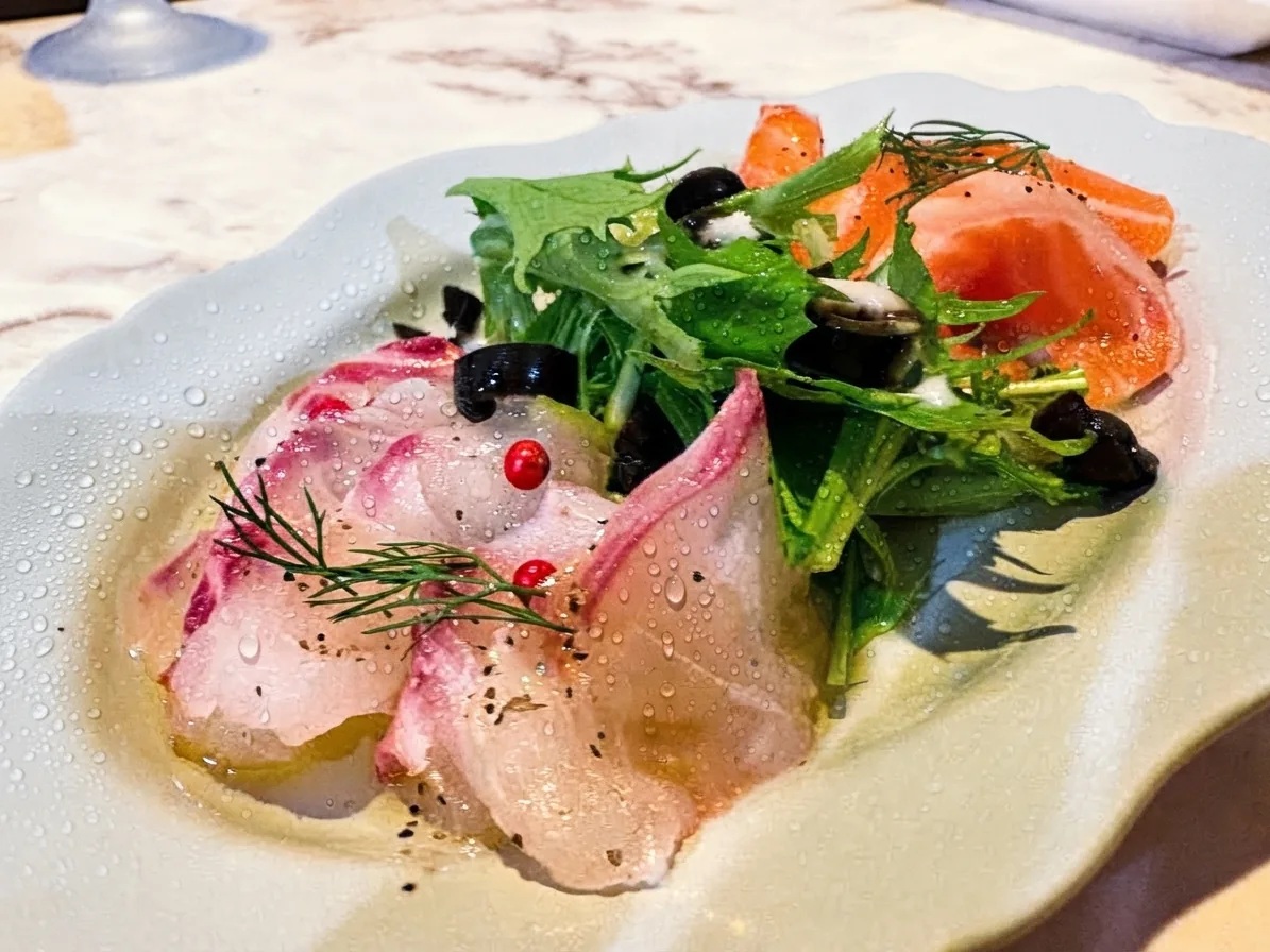 白い皿のカルパッチョ。白身魚とサーモン、ベビーリーフ、オリーブ、ピンクペッパー