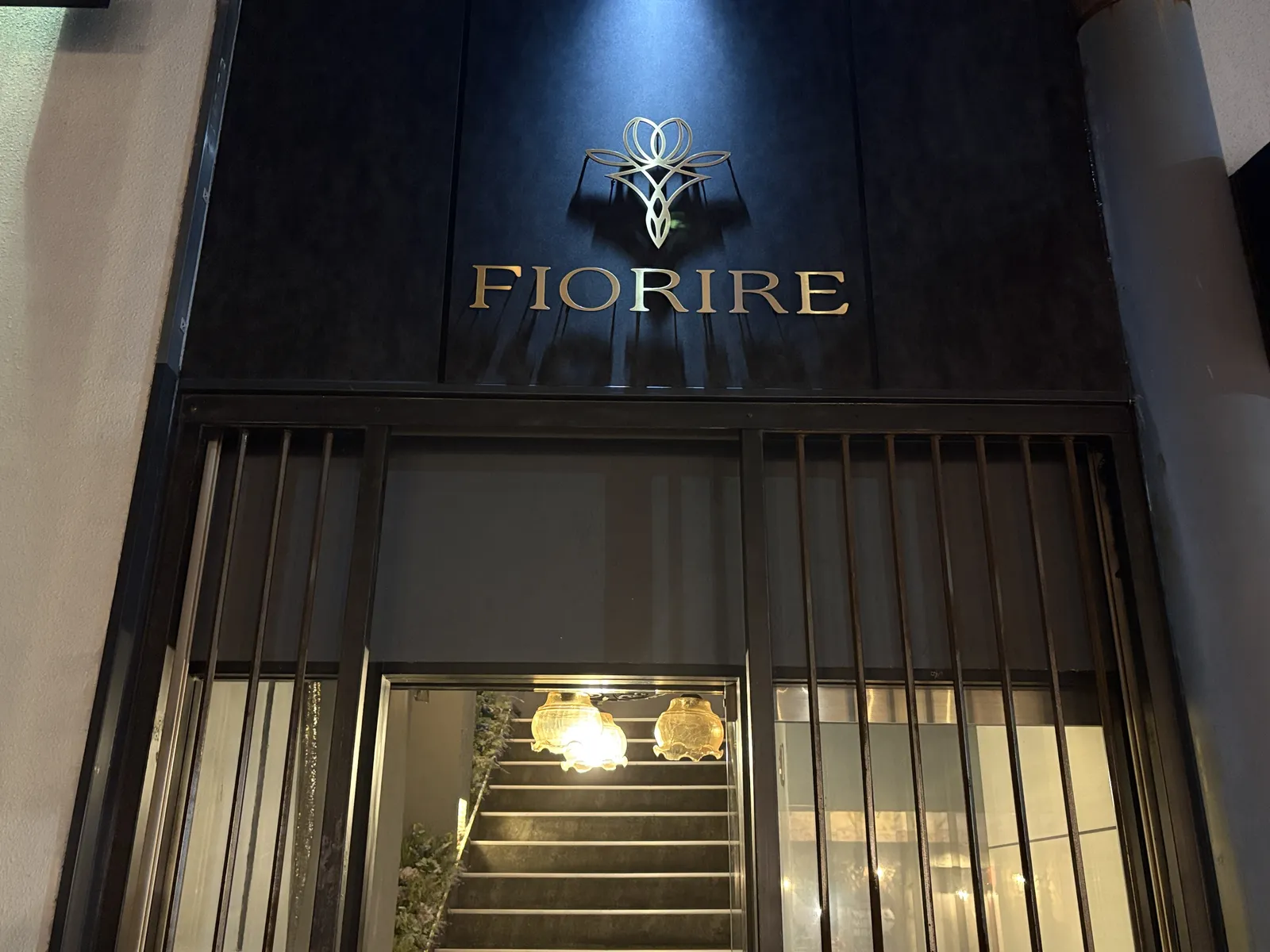 夜の入口。黒い壁の金色の「FIORIRE」のロゴが照明に浮かび、格子の奥に階段が見える