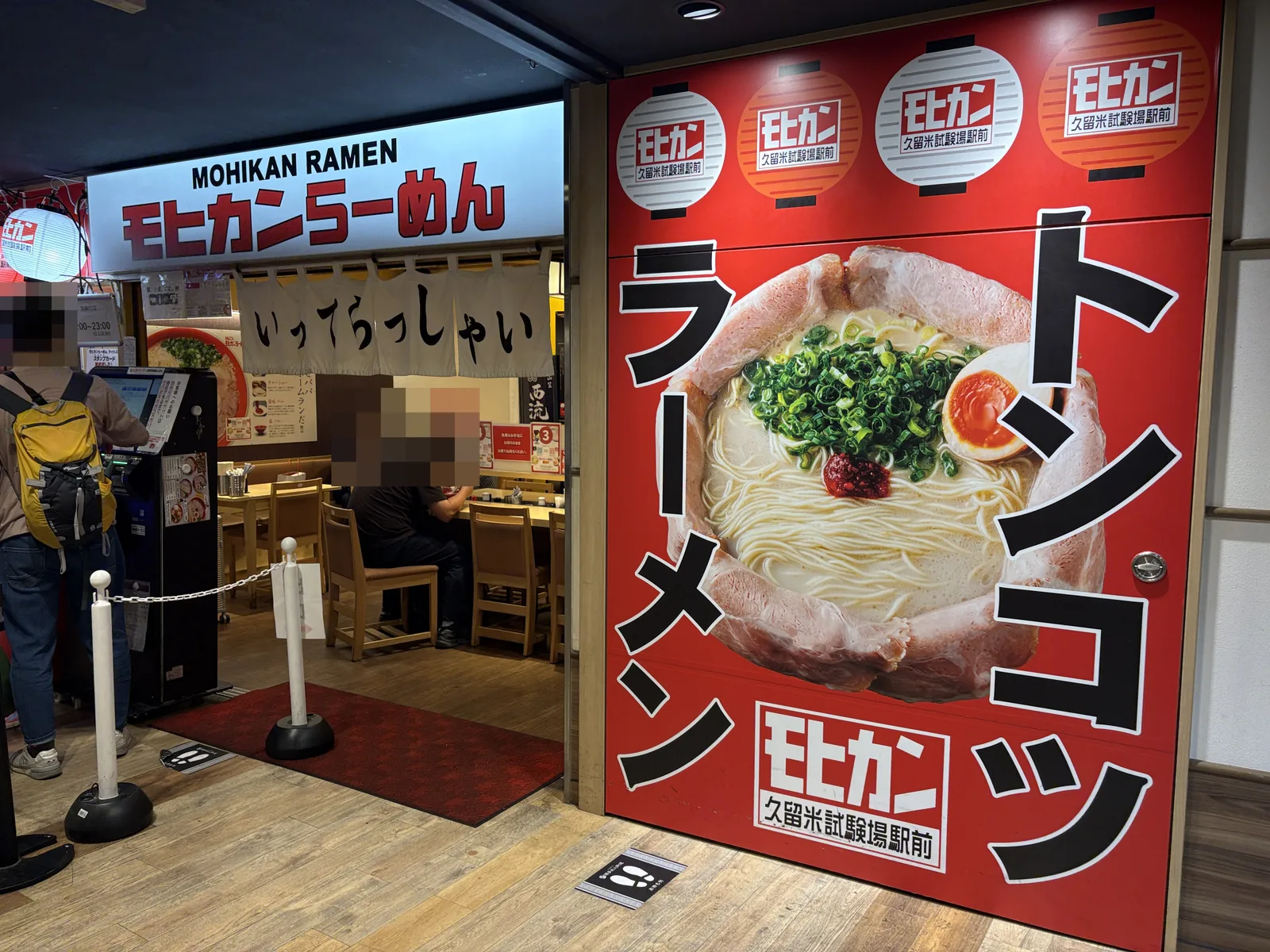 モヒカンらーめんの入口。券売機の前に客が並び、のれんに「いってらっしゃい」
