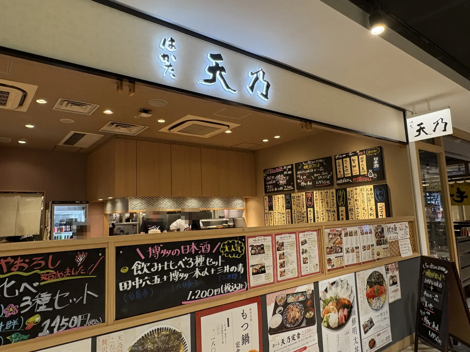はかた天乃の外観。カウンターの奥に厨房、店頭に「博多名物 もつ鍋定食」「海鮮明太丼」のポスターと日本酒の黒板