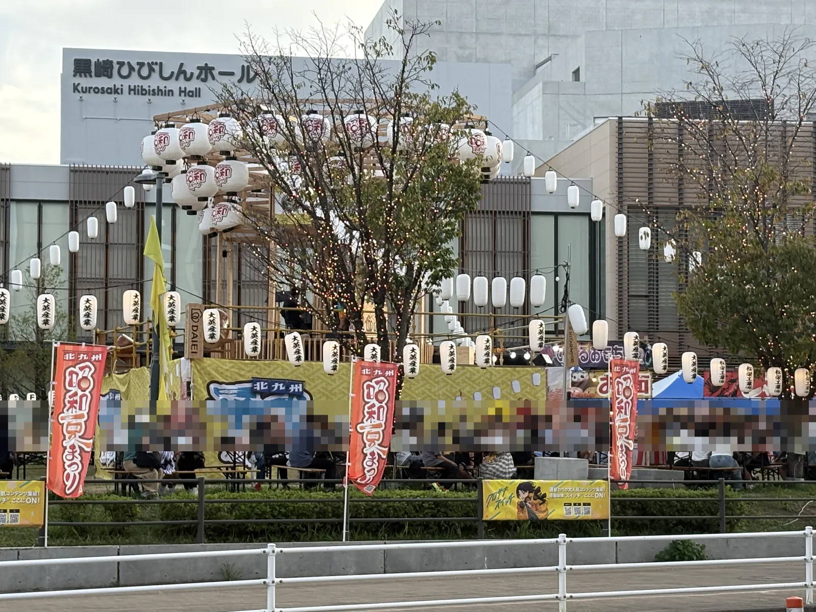 黒崎ひびしんホールの前に立つやぐら。提灯が並び、手前のテーブル席に人が座っている