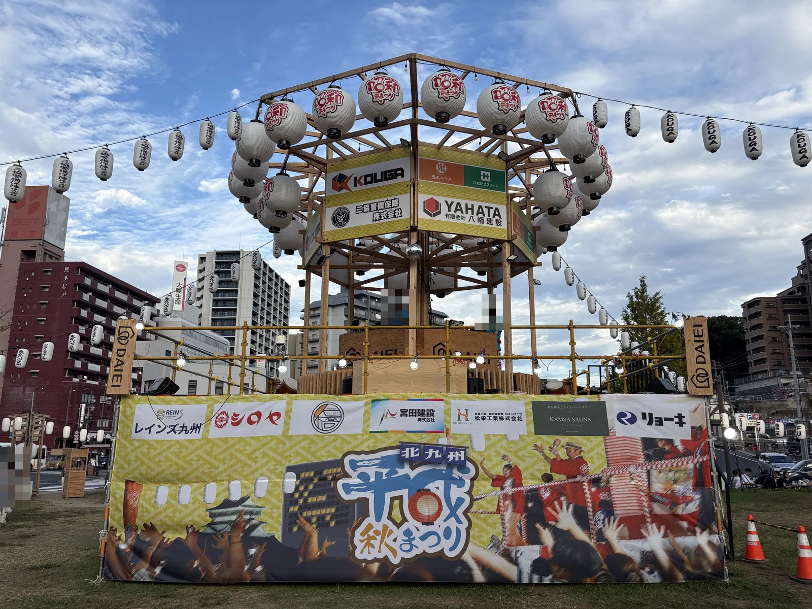 昼のやぐらの全景。木組みに提灯、協賛の看板、下に「北九州 平成秋まつり」の幕