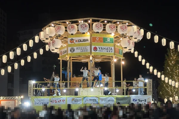 夜のやぐら。提灯と電飾に照らされ、上でダンサーが踊り、下に大勢の人が集まる