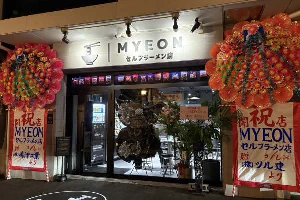 MYEON セルフラーメン店の外観。白い看板にラーメンどんぶりのロゴ、両脇に開店祝いの花輪