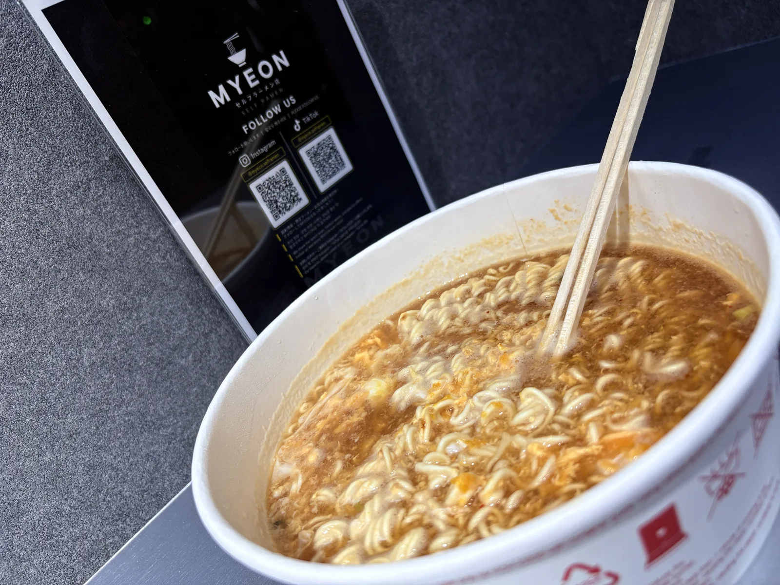 割り箸を入れたみそラーメン。奥にMYEONのInstagram・TikTokのQRコード