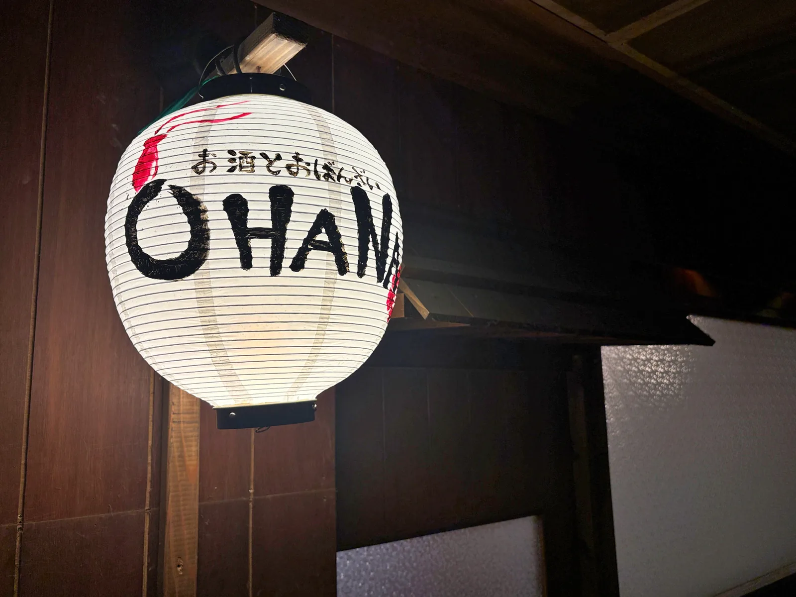 白い提灯に「お酒とおばんざい OHANA」の手書き文字。木の壁に灯りがともる