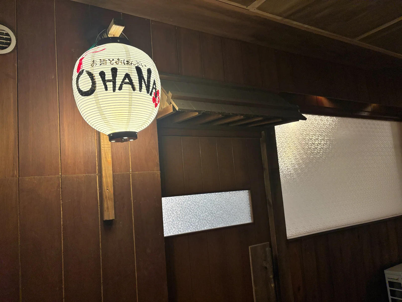 木の壁に掛かったOHANAの提灯と、すりガラスの入口