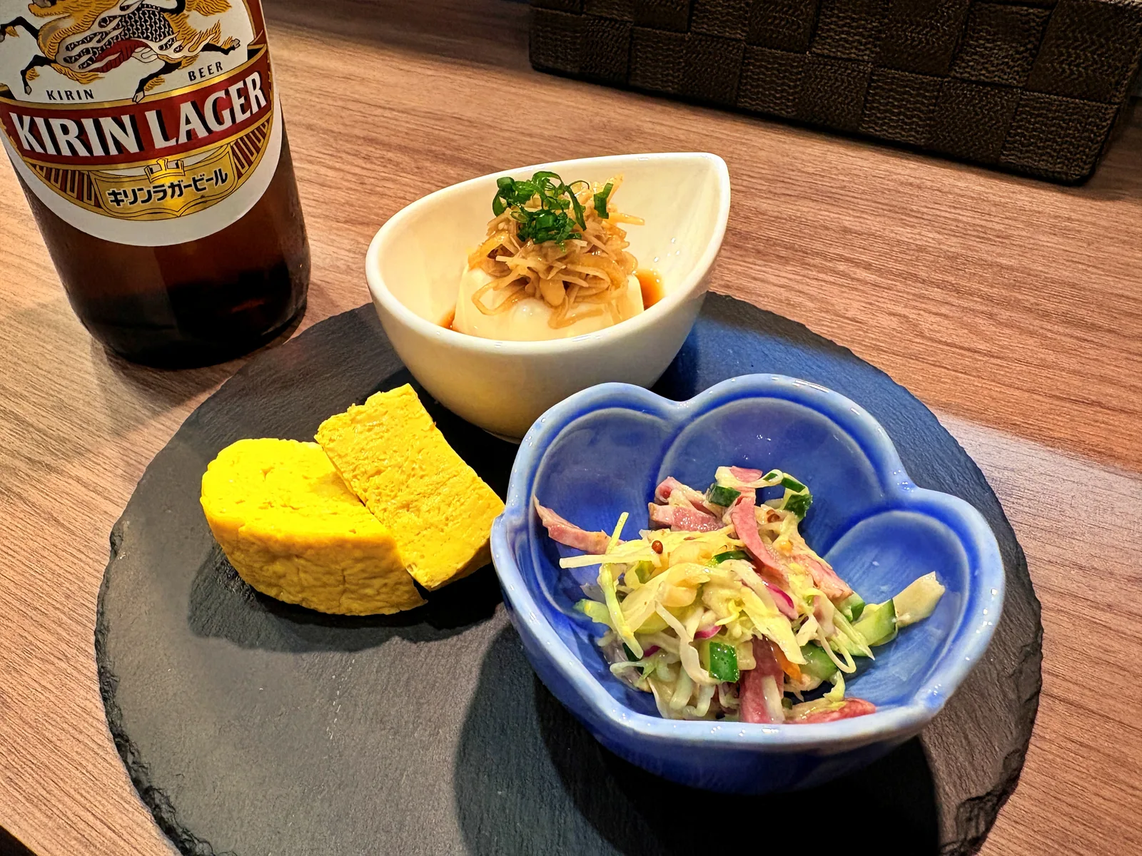 黒いスレートの皿に、厚焼きのだしまき玉子、豆腐の小鉢、ハム入りのサラダ。奥にキリンラガーの瓶