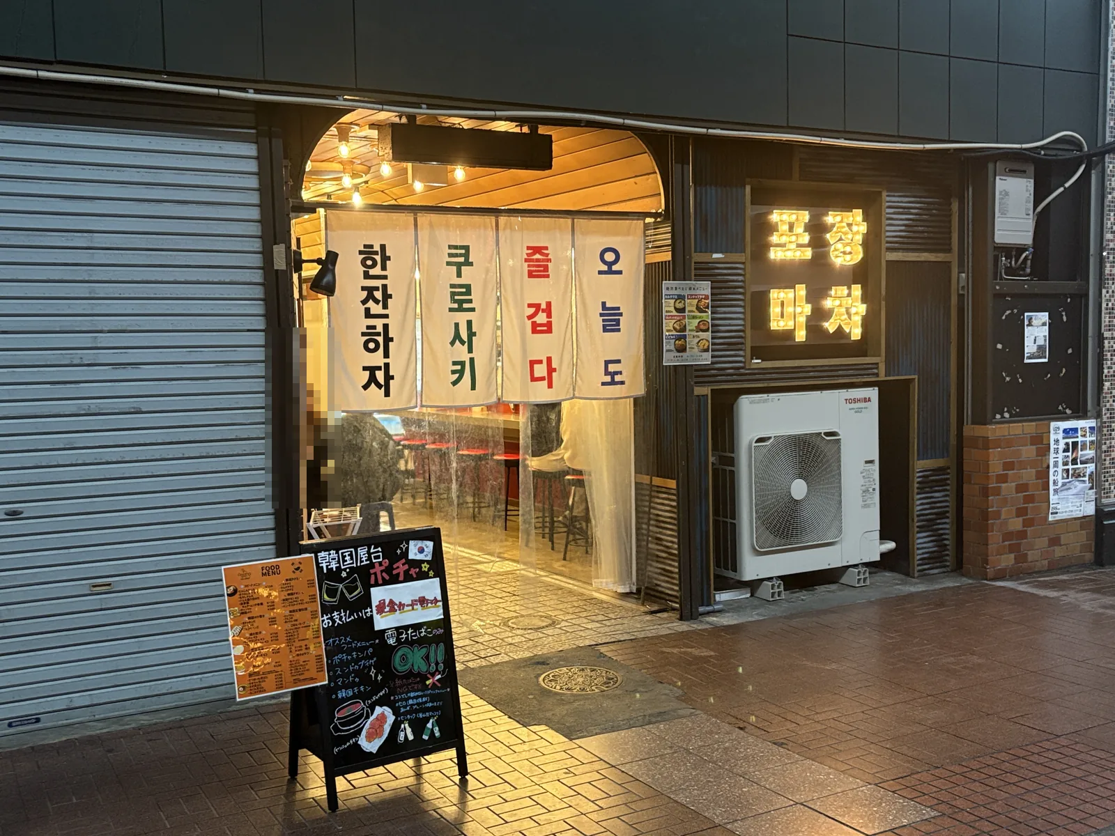 麻生ビル1階の韓国屋台ポチャの店頭。ハングルののれんが4枚下がり、手前に「韓国屋台ポチャ」の黒板が立つ