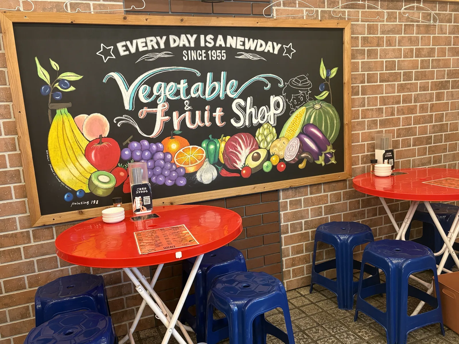 「Vegetable & Fruit Shop」と描かれた黒板アートの前に並ぶ赤いテーブル席