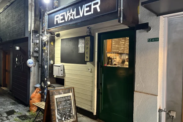 夜の外観。白く光る「REV☆LVER」の看板と緑色の扉。扉の小窓から店内の黒板メニューが見える