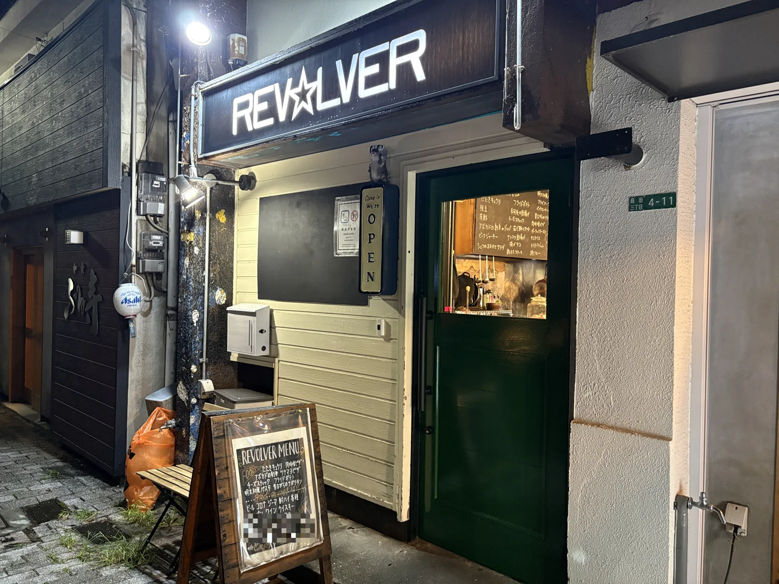 夜の外観。白く光る「REV☆LVER」の看板と緑色の扉。扉の小窓から店内の黒板メニューが見える