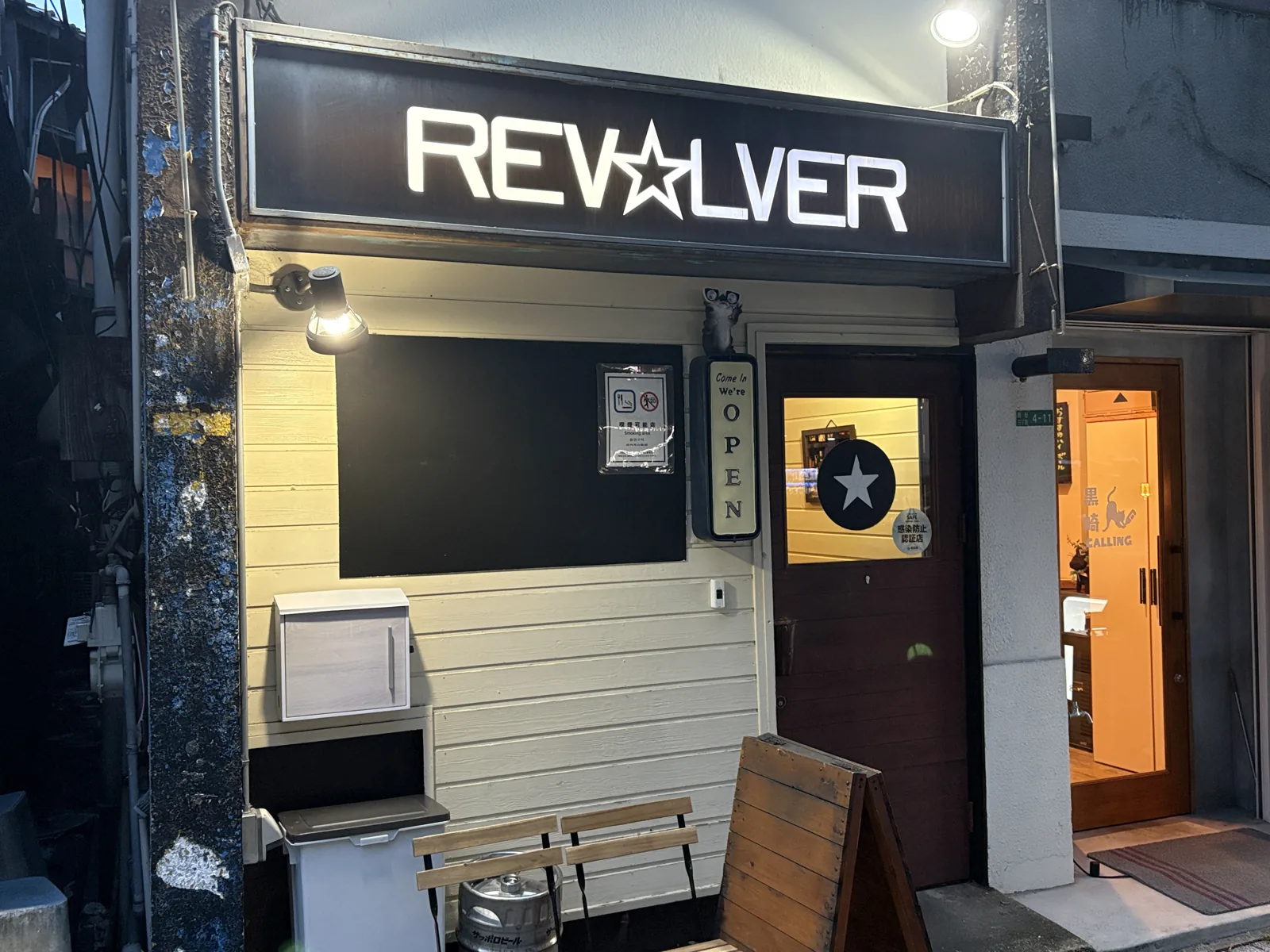 夕暮れどきの外観。白い板張りの壁に「Come In We're OPEN」の看板、店先にはベンチとサッポロビールの樽