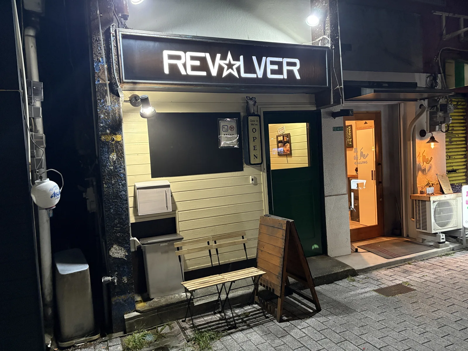 夜の外観。光る看板の下に緑色の扉とベンチ、右隣は黒崎CALLING