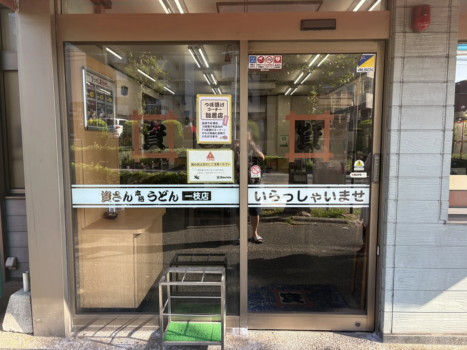 入口のガラス扉。「つぼ漬けコーナー設置店」の貼り紙と「資さん本格うどん 一枝店 いらっしゃいませ」の文字