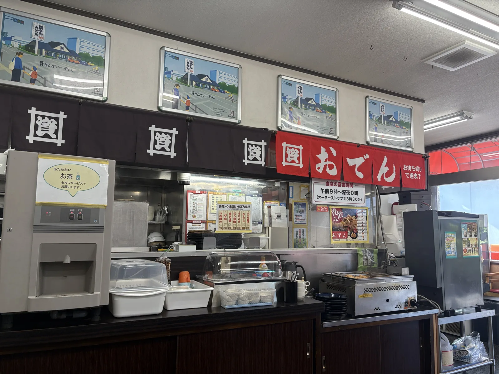 給茶機に「あたたかいお茶 セルフサービスでお願いします」。奥に薬味コーナー、右におでん