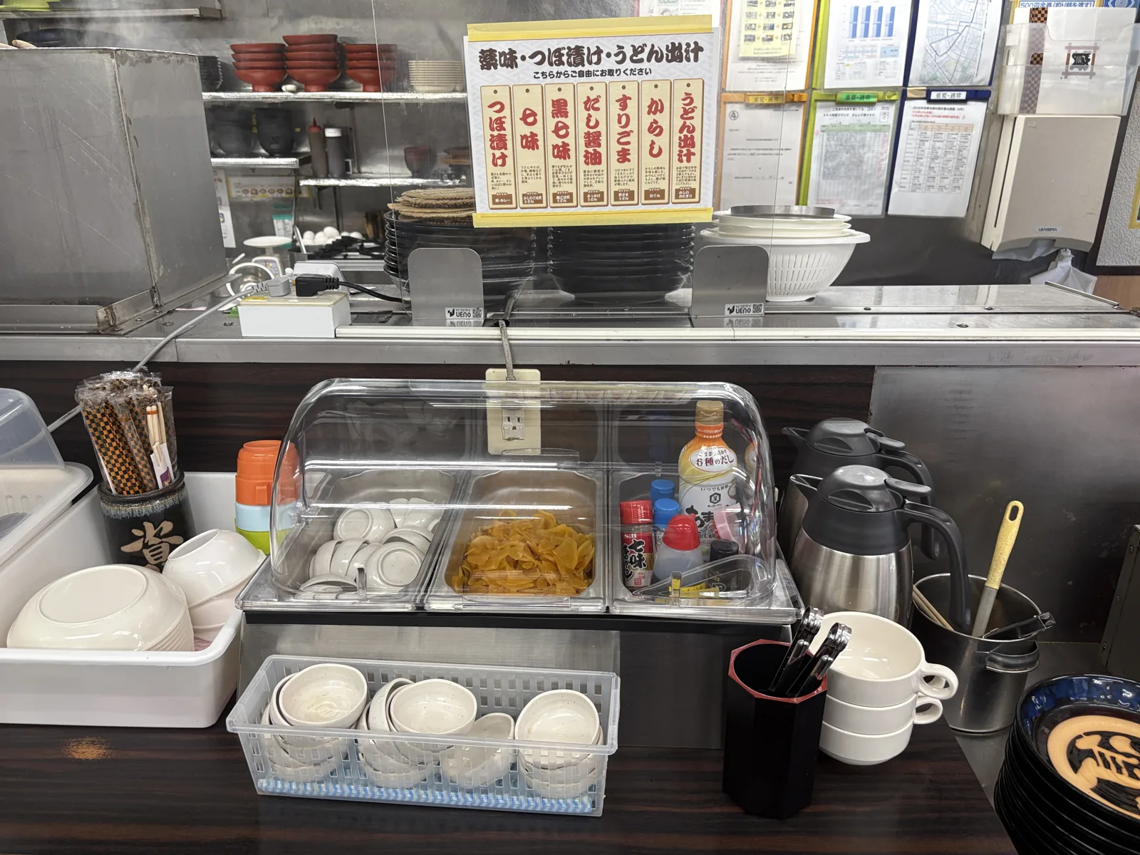 薬味コーナー。「薬味・つぼ漬け・うどん出汁」の看板に7種の札。クリアカバーの中につぼ漬けと小皿、調味料