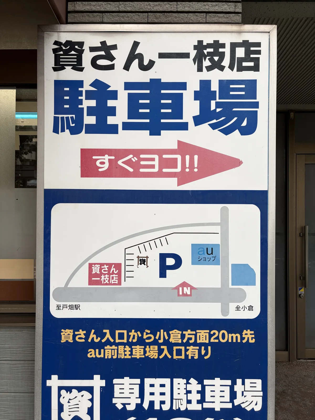 駐車場の看板。店の横にP、auショップ前に「IN」の矢印。「資さん入口から小倉方面20m先 au前駐車場入口有り」