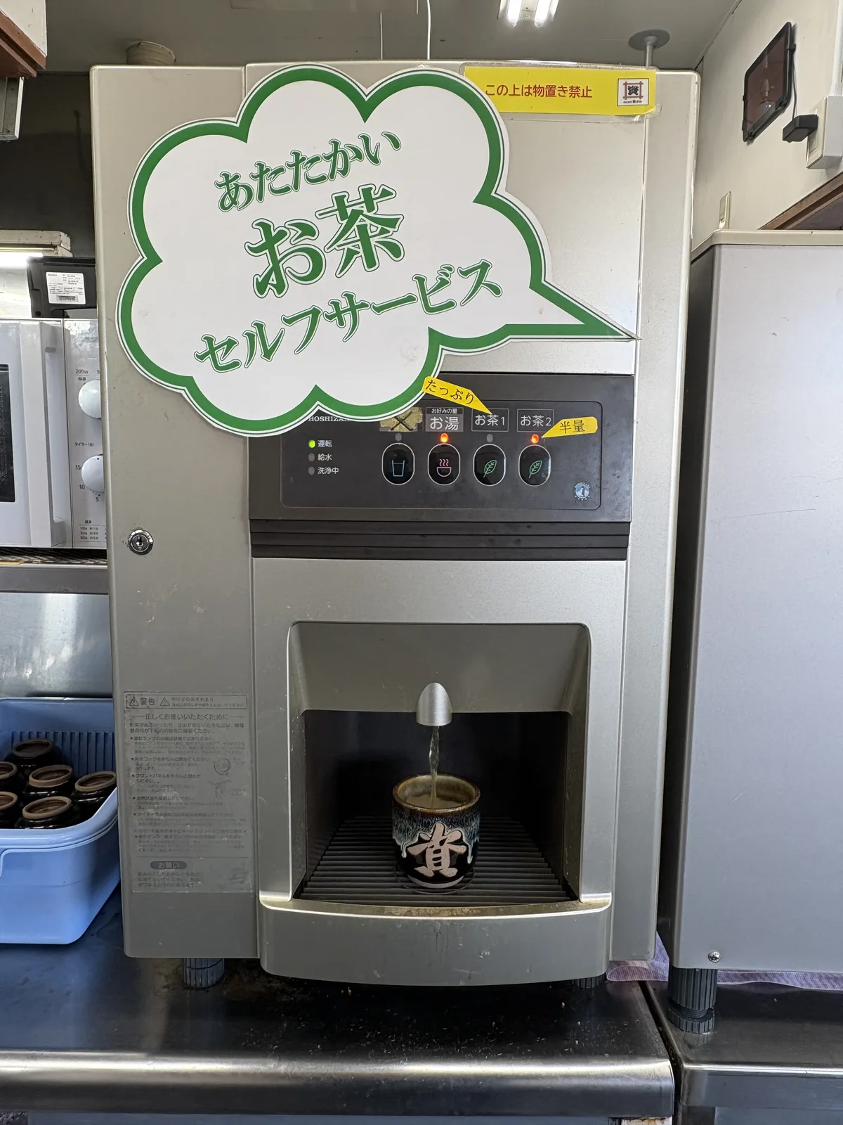 給湯器「あたたかいお茶 セルフサービス」。「たっぷり」「半量」のボタン表示。「資」の湯飲みにお茶が注がれている