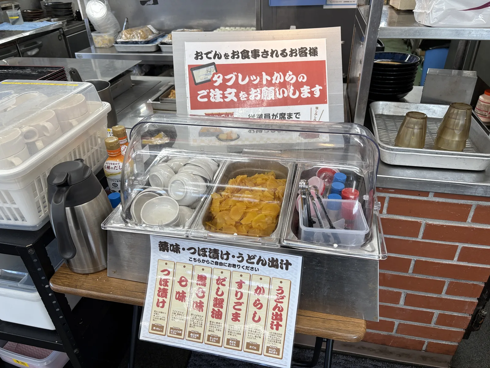 薬味コーナーのアップ。つぼ漬け、七味、黒七味、だし醤油、すりごま、からし、うどん出汁の札が並ぶ
