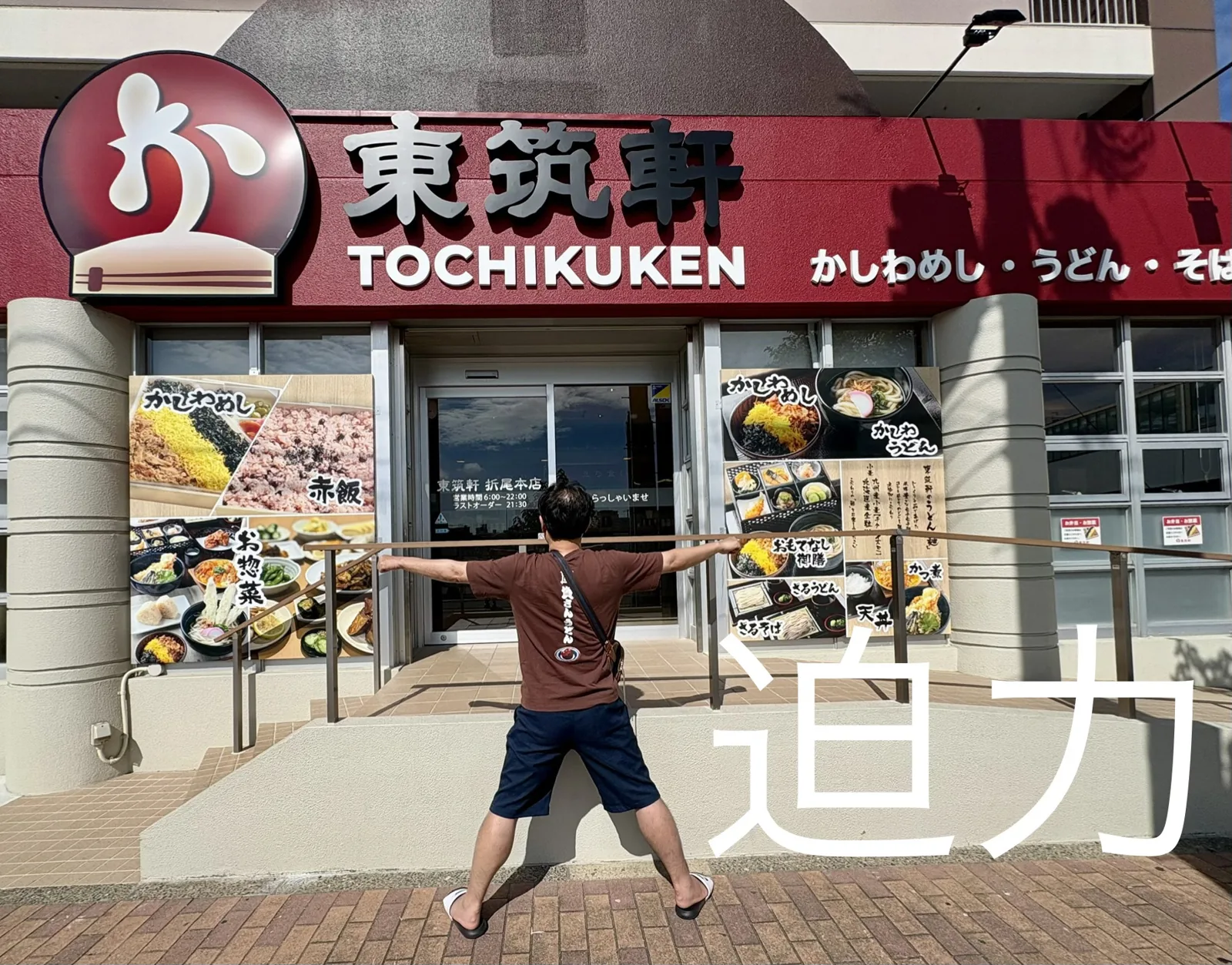 折尾本店の前で迫力ポーズ。赤い看板に「東筑軒 TOCHIKUKEN かしわめし・うどん・そば」