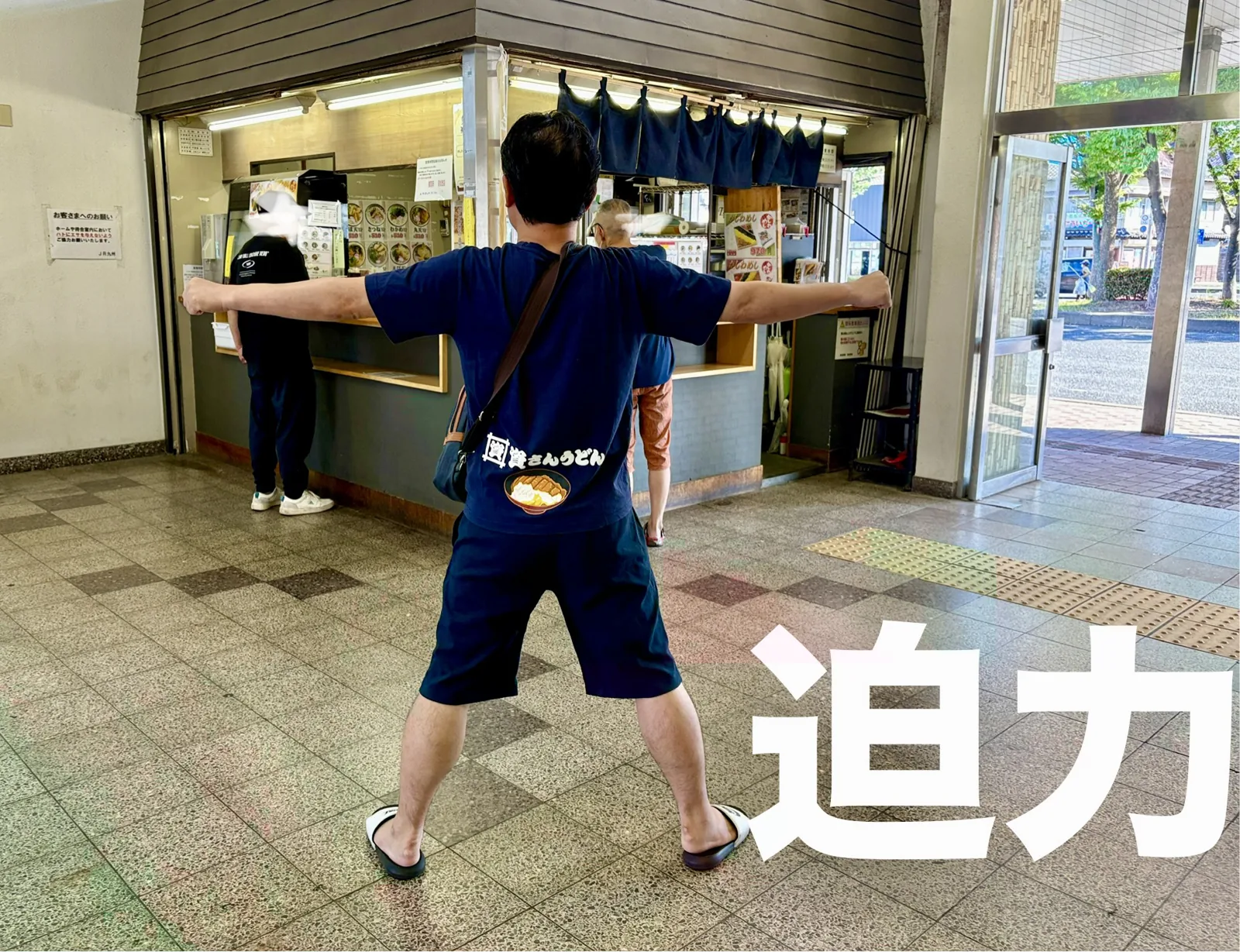 若松の店の前で迫力ポーズ。紺ののれんがかかる駅の立ち食いカウンター