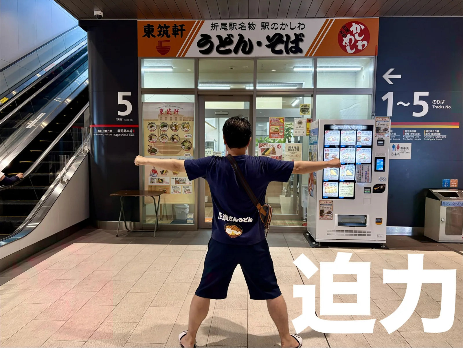 折尾駅の店の前で迫力ポーズ。「折尾駅名物 駅のかしわ うどん・そば」の看板、横に5番のりば
