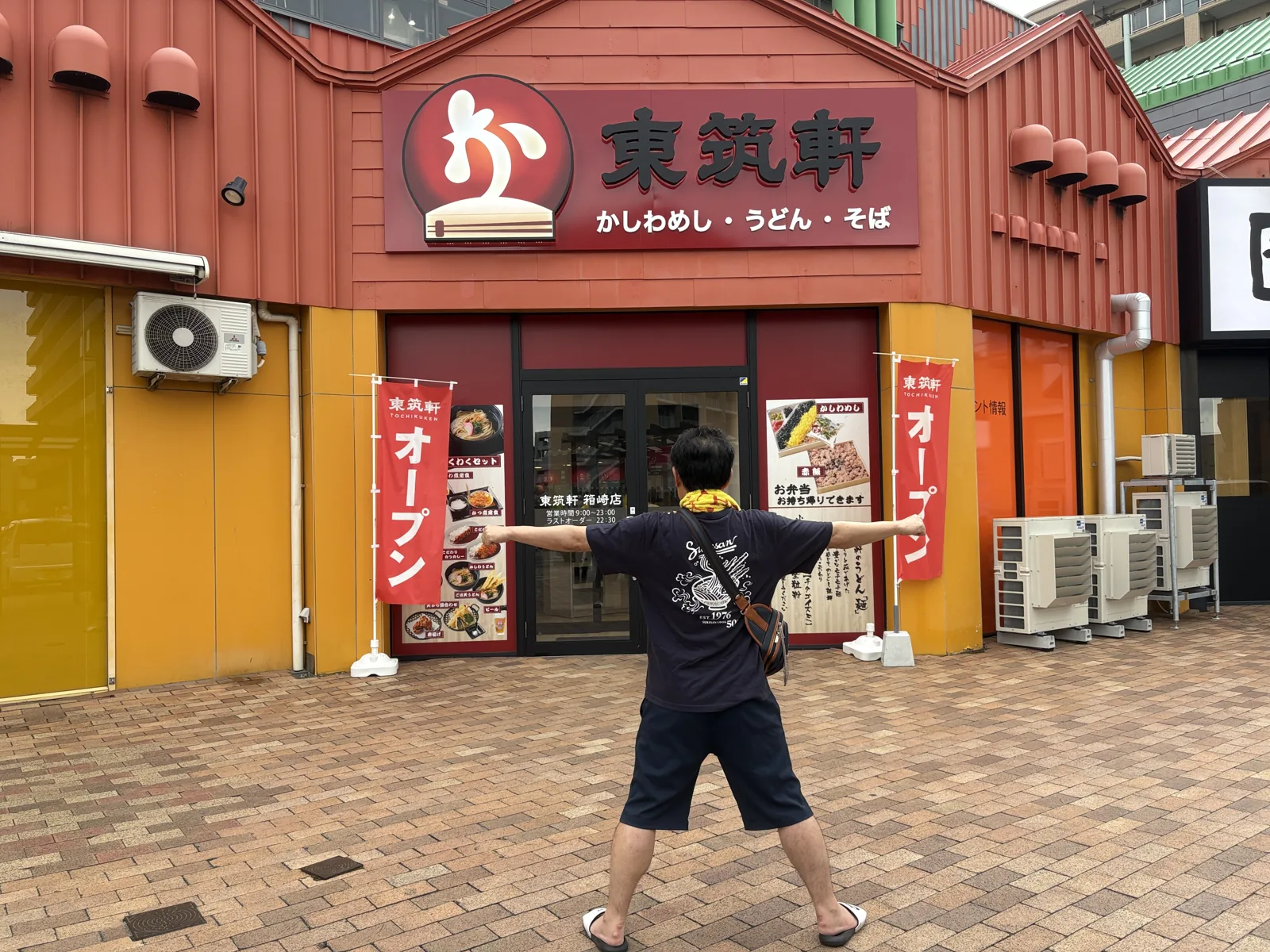 箱崎店の前で迫力ポーズ。赤い外壁に「オープン」ののぼり