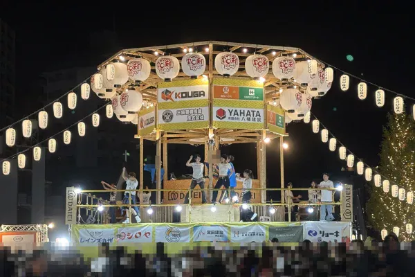 夜のやぐら。提灯と電飾に照らされ、上でダンサーが踊り、下に大勢の人が集まる