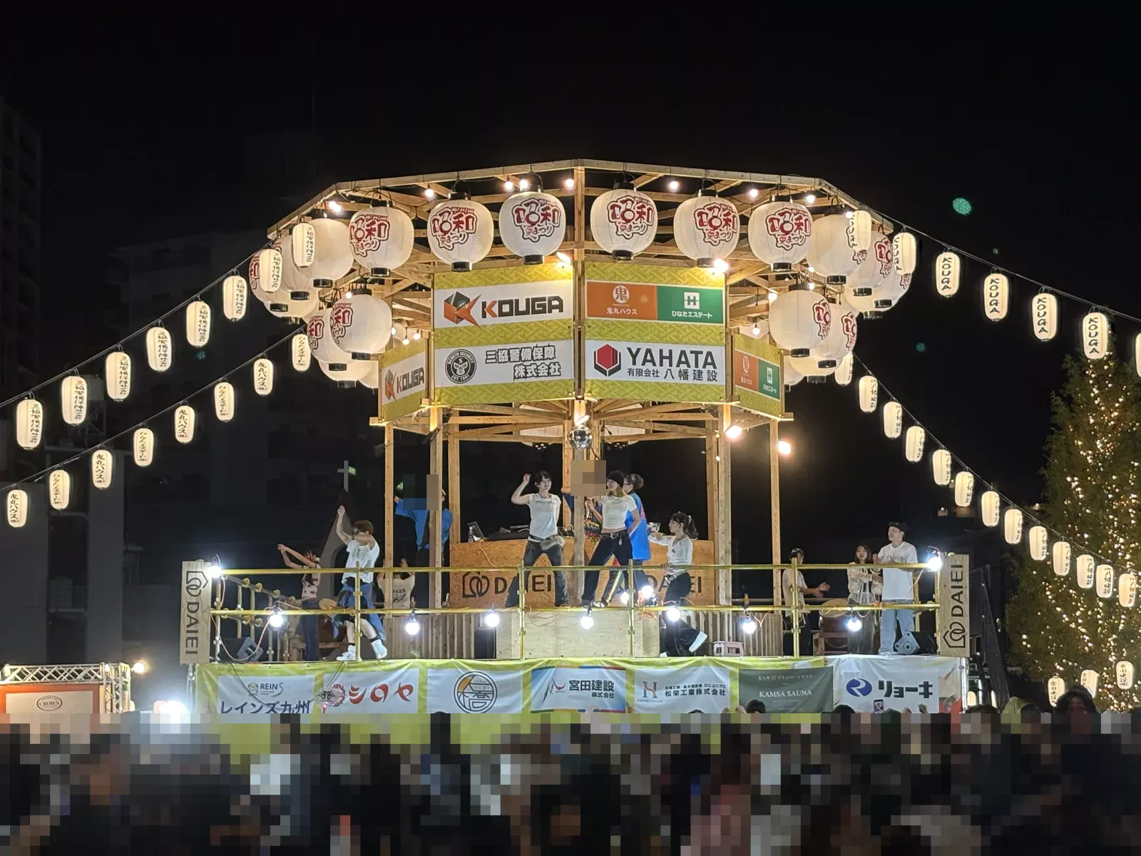 夜のやぐら。提灯と電飾に照らされ、上でダンサーが踊り、下に大勢の人が集まる