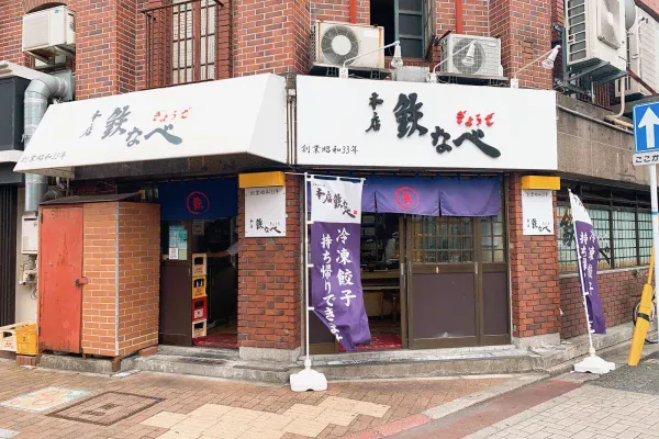 本店鉄なべ 黒崎本店の外観。白い看板に「本店 鉄なべ ぎょうざ」「創業昭和33年」、紫ののれんと「冷凍餃子 持ち帰りできます」ののぼり