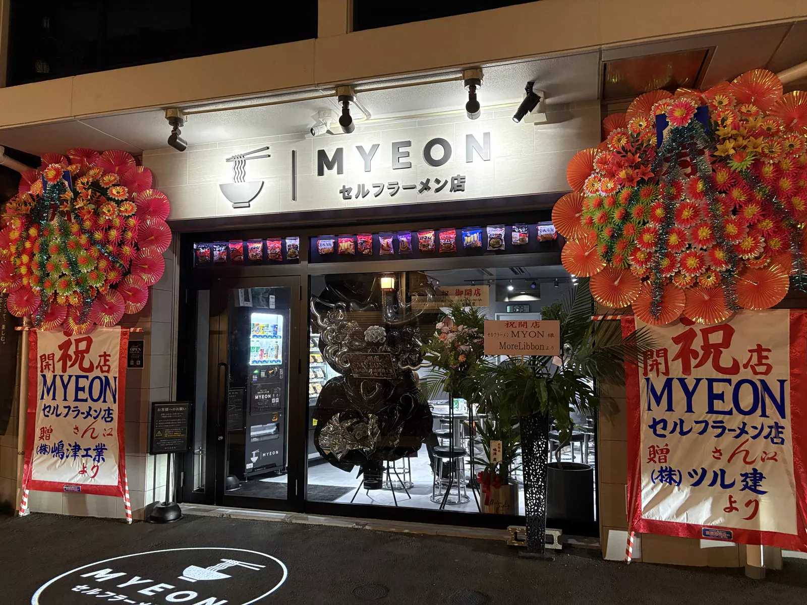 MYEON セルフラーメン店の外観。白い看板にラーメンどんぶりのロゴ、両脇に開店祝いの花輪