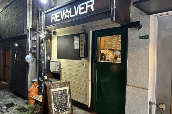 夜の外観。白く光る「REV☆LVER」の看板と緑色の扉。扉の小窓から店内の黒板メニューが見える