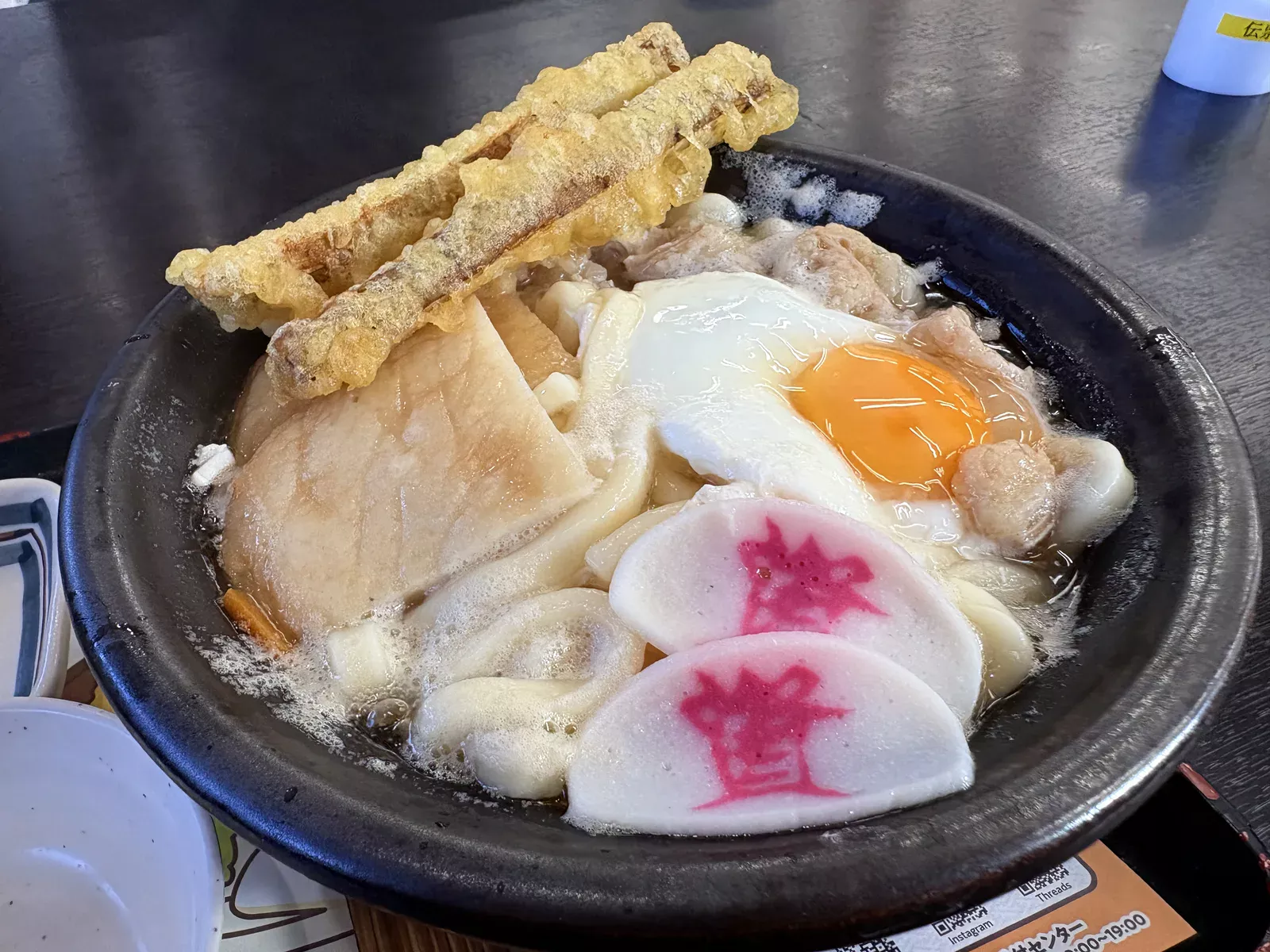 土鍋のもつ鍋うどん。ごぼう天2本、半熟の卵、「資」のかまぼこ2枚、もつがのる